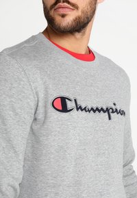 Grauer Sweatshirt mit Rundhalsausschnitt, versehen mit einem schwarzen, kursiven "Champion"-Logo und einem roten Akzent am Innenkragen. Weiche Textur.
