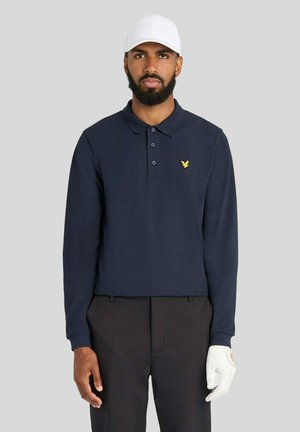 Mann mit Bart, der eine weiße Kappe, einen weißen Golfhandschuh, ein marineblaues Langarm-Poloshirt mit gelbem Vogel-Logo und schwarze Hose vor neutralem Hintergrund trägt.