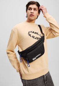Schwarze Nylon-Hüfttasche mit königsblauen Akzenten, ausgestattet mit einem Reißverschlussfach und Logo, getragen über einem beigen Sweatshirt.