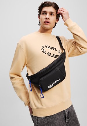 Schwarze Nylon-Hüfttasche mit königsblauen Akzenten, ausgestattet mit einem Reißverschlussfach und Logo, getragen über einem beigen Sweatshirt.