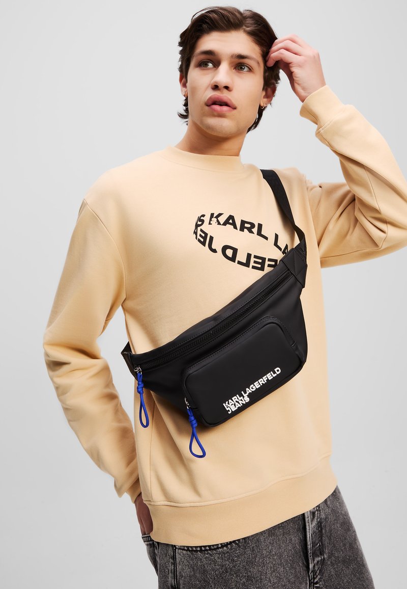 Schwarze Nylon-Hüfttasche mit königsblauen Akzenten, ausgestattet mit einem Reißverschlussfach und Logo, getragen über einem beigen Sweatshirt.