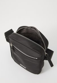 Sac bandoulière noir en tissu lisse, doté d'un compartiment principal zippé et d'une poche extérieure avec fermeture éclair argentée. Bandoulière réglable.