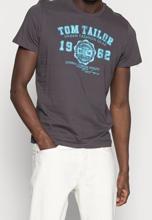 Homme portant un t-shirt Tom Tailor gris foncé avec texte imprimé en bleu et pantalon blanc, main dans la poche.
