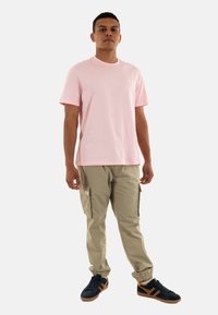 T-shirt in cotone rosa abbinato a pantaloni cargo leggeri color khaki con tasche. Sneakers nere con accenti arancioni, sfondo neutro.