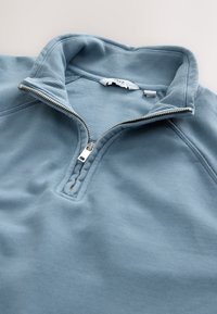 Helehall sinine neljandiklukuga dressipluus soonilise püstkraega ja osaliselt avatud hõbedase tõmblukuga ühevärvilisel taustal.