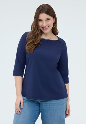 Usměvavá žena s dlouhými hnědými vlasy má na sobě tmavě modrý top s tříčtvrtečními rukávy a ozdobným výstřihem s cvočky, a světle modré džíny, stojí před neutrálním pozadím.