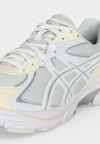 Hvite og beige treningssko med mesh-stoff, snøring og karakteristiske sidestiper på lys bakgrunn.