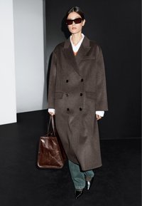 Brązowy wełniany płaszcz dwurzędowy z czarnymi guzikami, biała koszula, pomarańczowy akcent, zielone dżinsy, czarne szpilki oraz brązowa torebka.