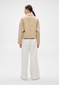 Béžová krátka bunda s golierom a širokými rukávmi, doplnená o biele nohavice s širokými nohavicami, ktoré majú jemnú textúru a zadné vrecká.