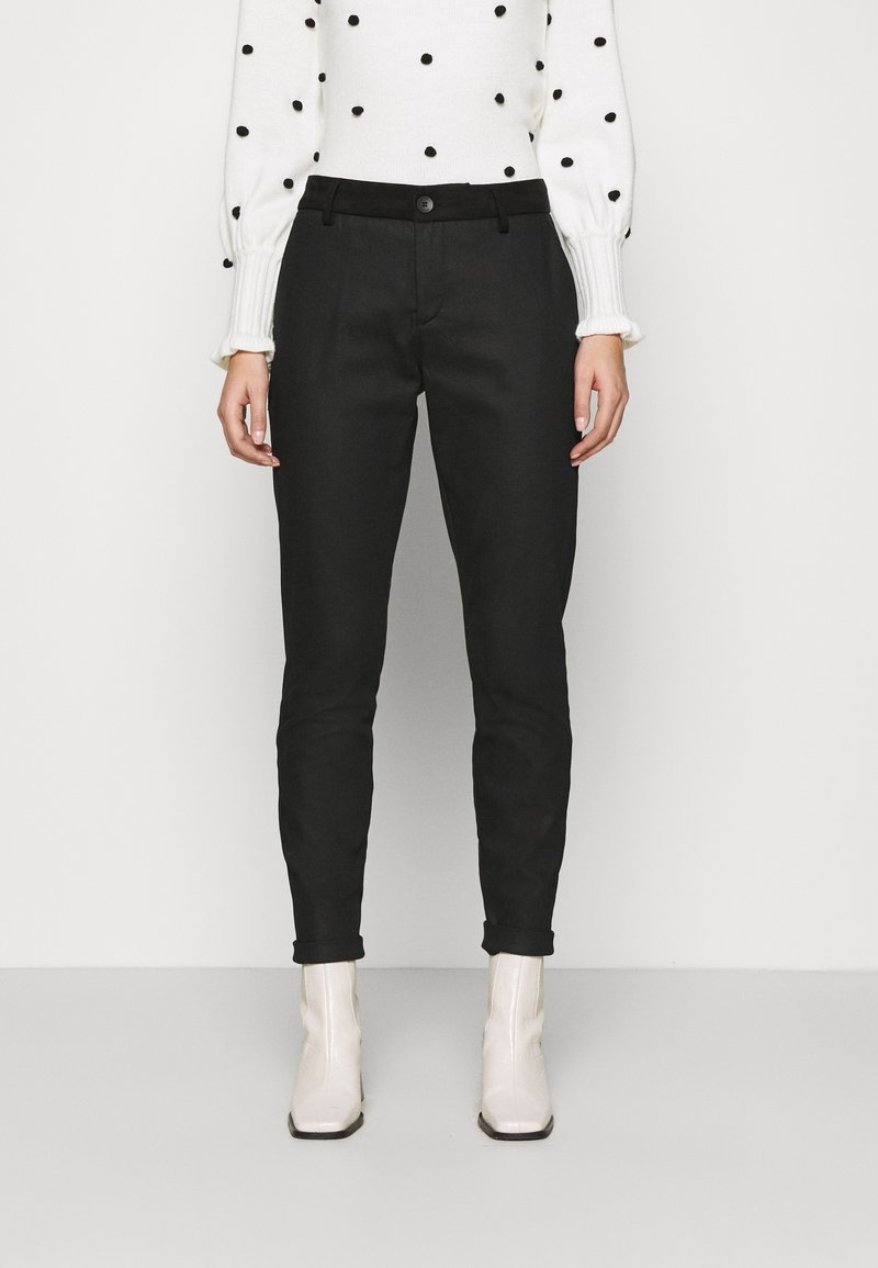 Czarne wąskie spodnie o gładkiej fakturze, z zapięciem na guziki z przodu i szlufkami na pasek, w zestawieniu z białym swetrem w groszki oraz jasnymi botkami za kostkę.