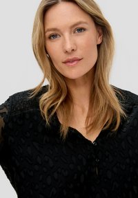 Zwarte blouse met een gestructureerd patroon en doorschijnende mouwen, voorzien van een knoopsluiting en een gesmokte schouderpartij.
