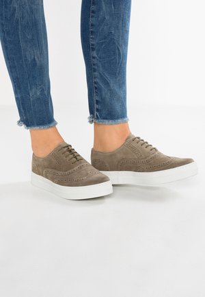 Taupekleurige suède vetersneakers met witte plateauzolen gedragen bij blauwe jeans met rafelige zoom op een witte vloer.