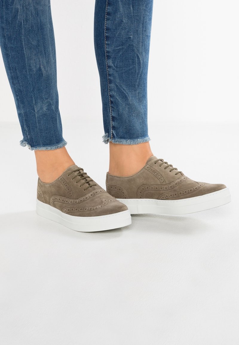 Baskets en daim taupe à lacets avec semelles compensées blanches portées avec un jean bleu à ourlet effiloché sur un sol blanc.
