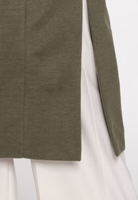 Olijfgroene gestructureerde jas met een rechte snit en open voorkant, gedragen over witte broek, wat een kleurcontrast benadrukt.