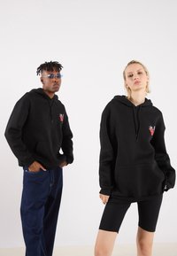 Black hoodies with front pockets, featuring a red graphic detail. The male model wears blue jeans, and the female model wears black bike shorts.