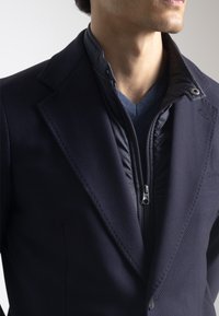 Blazer azul marino texturizado con un forro interior extraíble con cremallera y un suéter de cuello en V debajo, con una tela suave y un diseño ajustado.