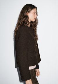 Jonge vrouw met lang krullend haar draagt een donkerbruine knoopjasje, lichte top en bruine broek, terwijl ze zijwaarts staat tegen een effen lichte achtergrond.