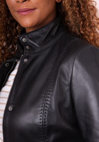 Black leather jacket with a high collar, metal snaps, and visible stitching. Soft texture and fitted design, featuring a zip closure.
