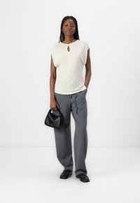 Haut beige clair à manches courtes avec un détail en forme de clé, associé à un pantalon gris ample et un petit sac à main noir. Texture de tissu lisse.