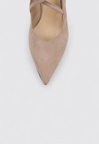 Zapato de ante beige de punta afilada con un corte en forma de V y tiras entrecruzadas. La suela tiene un borde negro. Textura suave en general.