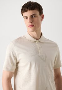 Smėlinė polo marška su trumpomis rankovėmis, puošta subtiliu gėlių raštu. Su apykakle, kurią puošia du mygtukai ties kaklu. Minkšta tekstūra.