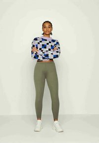 Suéter de malha multicolorido com um padrão de quadrados roxo, azul e preto, combinado com leggings justas verde oliva e ténis brancos.
