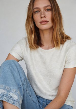 Young woman with shoulder-length light brown hair, wearing a textured white short-sleeve top and blue embroidered wide-leg jeans, seated calmly.