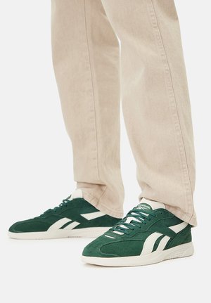 Zielone zamszowe trampki z białymi akcentami, o niskim kroju i wiązane na sznurowadła, zestawione z jasnobeżowymi spodniami.