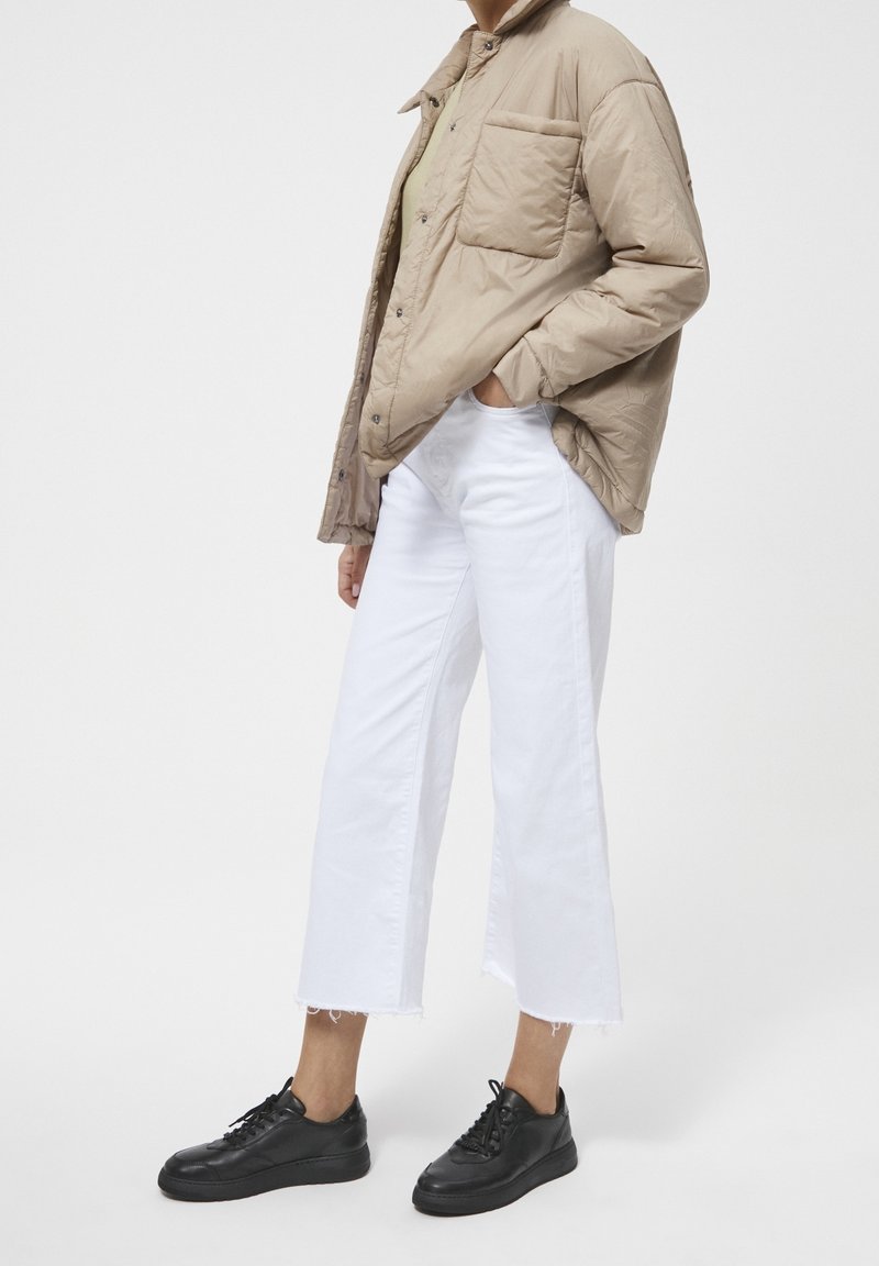 Personne portant une veste matelassée beige, un jean blanc court à jambes larges avec des ourlets bruts, et des baskets noires à lacets, debout de côté.