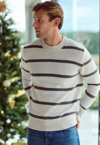 Light grey knit sweater with dark grey horizontal stripes, round neckline, and ribbed cuffs. Worn with blue jeans, against a festive backdrop.