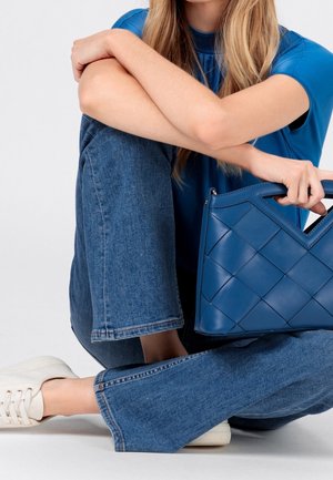 Blå quiltad handväska i slätt läder med geometrisk design. Har ett handtag på toppen och står bredvid vita sneakers på jeansklädda ben.