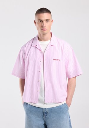 Jonge man met kort haar, die een roze-wit gestreept overhemd met korte mouwen draagt over een wit t-shirt en lichtblauwe spijkerbroek, handen in de zakken.