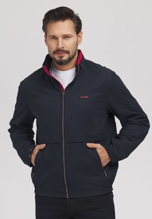 Mężczyzna o krótkich brązowych włosach i brodzie, ubrany w granatową kurtkę na suwak z czerwonym podszyciem, ręce w kieszeniach, neutralne szare tło.