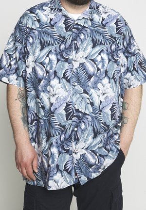 Mand iført en blå og hvid kortærmet skjorte med knapper og tropisk blad- og blomsterprint, hænder delvist synlige med tatoveringer.