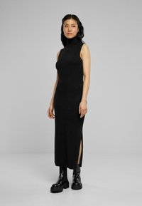 Robe noire sans manches, à col haut, en tissu doux, avec des fentes latérales à l'ourlet, portée avec des bottes noires imposantes.