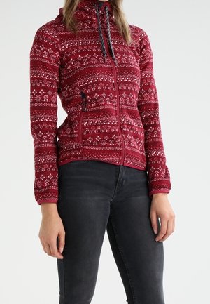 Vrouw draagt een nauwsluitend rood fleecejack met ritssluiting, witte geometrische patronen en zwarte trekkoorden, gecombineerd met donkergrijze nauwsluitende spijkerbroek.