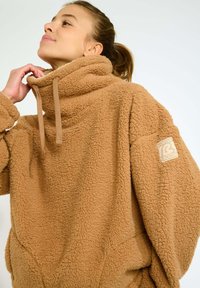 Felpa in pile marrone con colletto alto, superficie testurizzata e toppa con logo sulla manica. Ha una vestibilità ampia e un dettaglio con cordoncino.