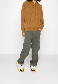 Sudadera marrón con un patrón, con cordones y bolsillos laterales, combinada con pantalones cargo verde oliva con grandes bolsillos y zapatillas de colores.