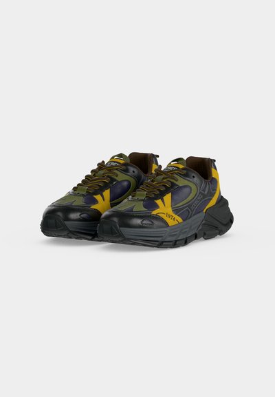 Paire de baskets épaisses noires, jaunes, vert olive et bleu marine avec semelles épaisses et design à lacets, inclinées vers la droite sur fond blanc.