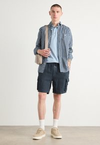 Tenue décontractée comprenant une chemise à carreaux bleus, un polo bleu clair, un short cargo noir et des baskets beiges avec des chaussettes blanches. Petit sac en bandoulière.