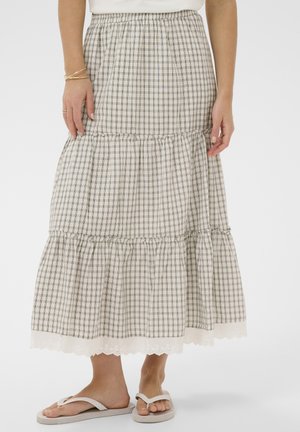 Frau trägt einen beige-weiß karierten mehrlagigen Rock mit Spitzensaum, weiße Flip-Flops sowie goldene Armbänder und Ringe an den Händen.