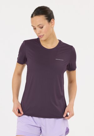 Junge Frau trägt ein dunkelviolettes Endurance-T-Shirt und hellviolette Shorts, schaut nach unten und hält den Saum des T-Shirts mit beiden Händen.