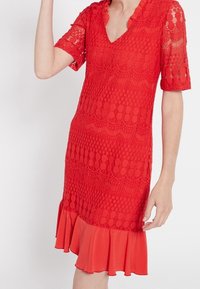 Červené krajkové šaty s krátkými rukávy, výstřihem do V a vlnitým lemem. Obsahují texturované vzory a vyztuženou volánkovou sukni na spodní části.