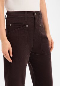 Pantalon en velours côtelé brun foncé avec une finition texturée, doté de deux poches avant et d'une fermeture par bouton. Coutures détaillées et accents latéraux visibles.