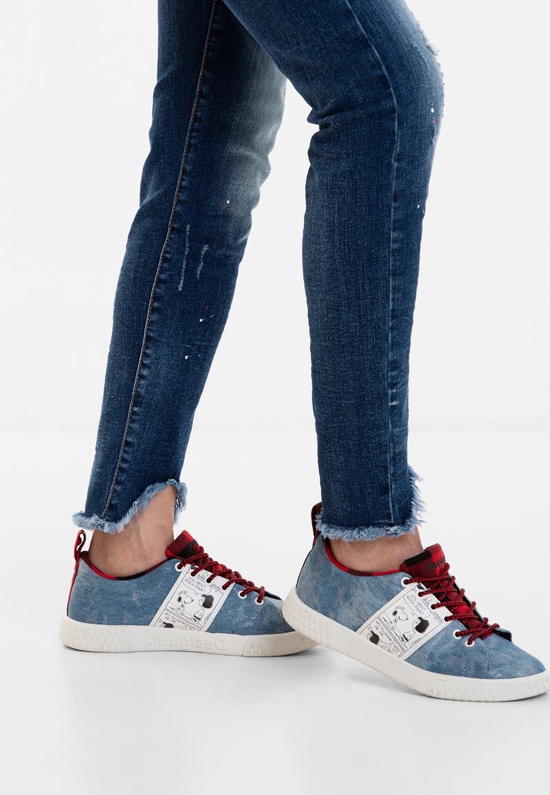Valkoiset denim-villakengät, joissa on vaaleansininen päällinen, punaiset nauhat ja kuvioitu valkoinen sivupaneeli. Korostettu teksturoidulla valkoisella pohjalla.