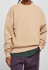 Beiger Pullover mit Rundhalsausschnitt und überschnittenen Schultern, aus weichem Stoff, kombiniert mit einem grünen Hemd darunter und lila Hose.