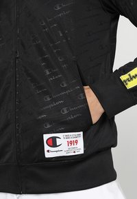 Svart zip-up jacka med subtilt "Champion" logomönster. Har en framficka och en märkt tagg med "1919" på nedre vänstra sidan.
