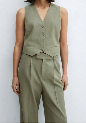 Donna che indossa un gilet verde oliva senza maniche con bottoni e pantaloni plissettati a vita alta abbinati su uno sfondo neutro.