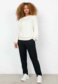 Ljus krämfärgad sweatshirt med långa ärmar, kombinerad med svarta joggers. Slät tygtextur, med sidofickor och elastiska ärmslut. Vita sneakers.