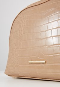 Borsa beige in ecopelle con texture effetto coccodrillo, forma arrotondata e targhetta del marchio color oro. Superficie liscia con finitura lucida.